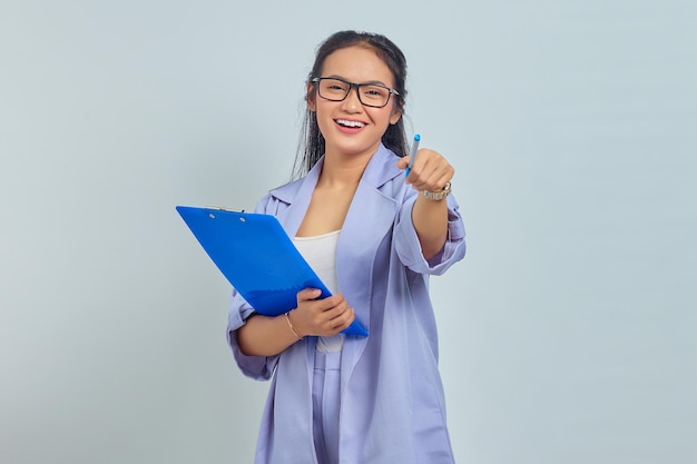 Retrato de una hermosa joven mujer de negocios asiática con traje de pie sosteniendo una carpeta de documentos y señalando con un bolígrafo mirando a la cámara con cara sonriente aislada en un fondo morado