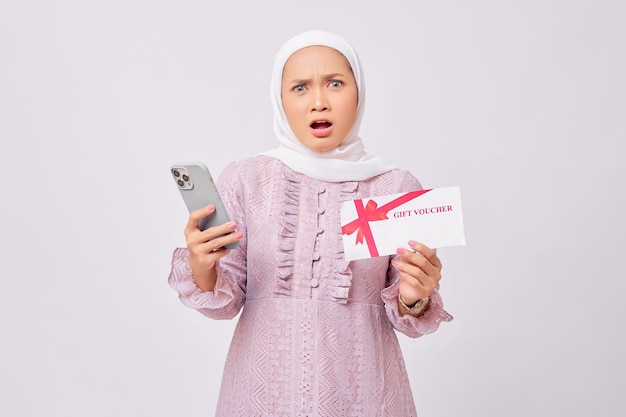 Retrato de una hermosa joven musulmana asiática sorprendida con hiyab y vestido morado con un certificado de vale de regalo y un teléfono móvil aislado en un estudio blanco