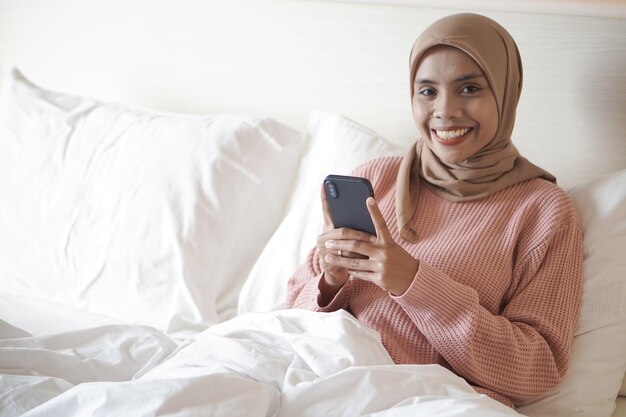 Retrato de una hermosa joven musulmana asiática usando hijab en la cama sosteniendo un teléfono móvil