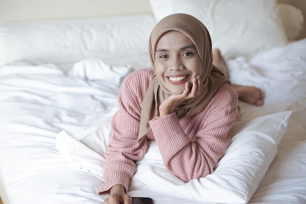 Retrato de una hermosa joven musulmana asiática usando hijab en la cama sosteniendo un teléfono móvil