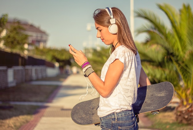 Foto retrato de hermosa joven con patineta y auriculares escuchando música en su teléfono inteligente al aire libre. edición en tonos cálidos.