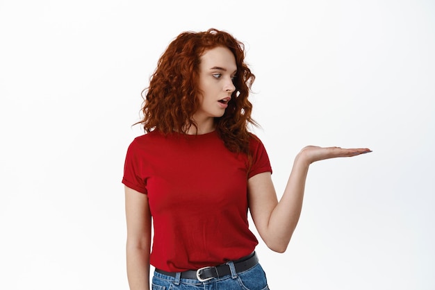 Retrato de una hermosa joven pelirroja con peinado rizado mirando su palma como si estuviera sosteniendo un objeto mirando a la mano con un producto de fondo blanco