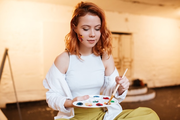 Foto retrato de la hermosa joven pintora pelirroja con pinturas al óleo en la cara en el taller del artista.