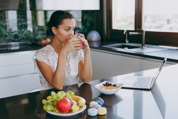 El retrato de la hermosa joven que trabaja con la computadora portátil mientras desayuna con cereales y leche y bebe jugo de naranja