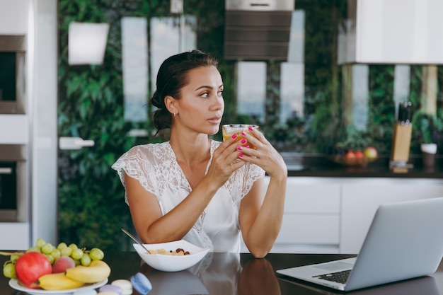 El retrato de la hermosa joven que trabaja con la computadora portátil mientras desayuna con cereales y leche y bebe jugo de naranja