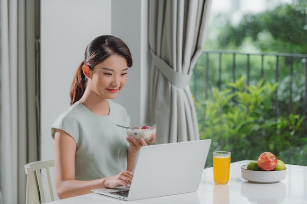 Retrato de una hermosa joven que trabaja con una laptop mientras come en casa