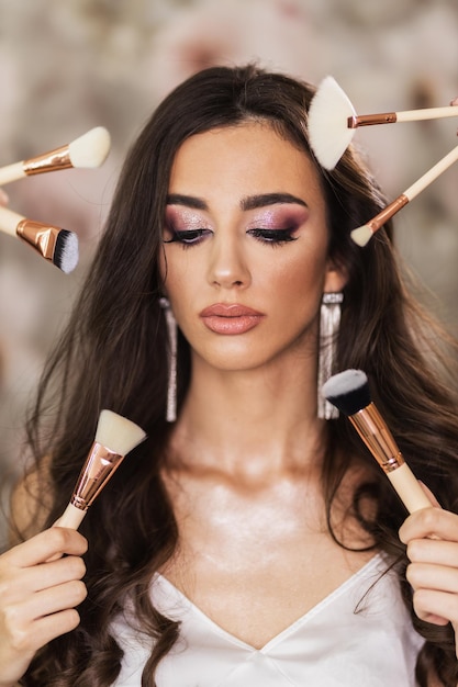 Un retrato de una hermosa joven rodeada de pinceles de maquillaje que presenta un bonito maquillaje en su rostro.