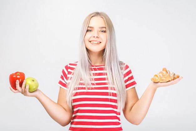 Retrato de hermosa joven rubia eligiendo entre un alimento saludable y no saludable. Aislado sobre fondo blanco.