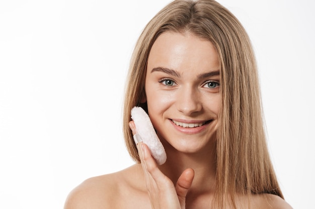 Retrato de hermosa joven rubia semidesnuda aplicando maquillaje con esponja en polvo aislado en blanco