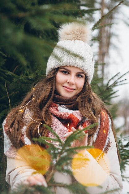 Retrato de una hermosa joven con un sombrero entre árboles de Navidad de invierno con luces. Un parque con árboles de Navidad al fondo. Humor navideño. Tintes.