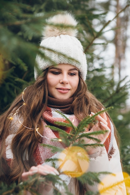 Retrato de una hermosa joven con un sombrero entre árboles de Navidad de invierno con luces. Un parque con árboles de Navidad al fondo. Humor navideño. Tintes.