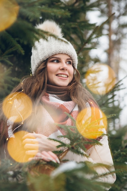 Retrato de una hermosa joven con un sombrero entre árboles de Navidad de invierno con luces. Un parque con árboles de Navidad al fondo. Humor navideño. Tintes.