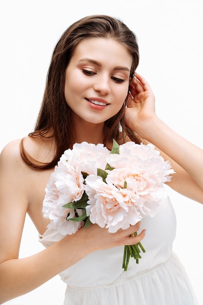 Retrato hermosa joven sonriendo y posando con flores artificiales en la pared blanca en vestido blanco