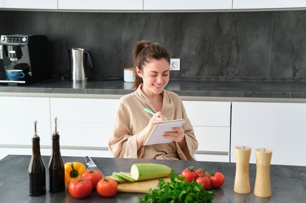 Retrato de una hermosa joven sonriente haciendo una lista de comidas escribiendo una receta sentada en el