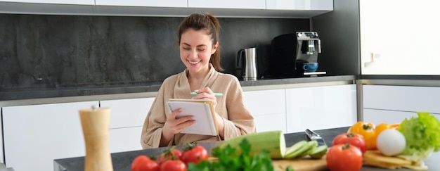 Retrato de una hermosa joven sonriente haciendo una lista de comidas escribiendo una receta sentada en el