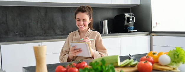 Foto retrato de una hermosa joven sonriente haciendo una lista de comidas escribiendo una receta sentada en el