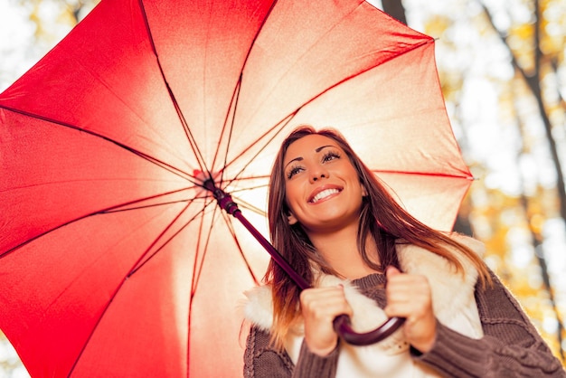 Retrato de una hermosa joven sonriente con paraguas rojo en un bosque soleado en colores otoñales.