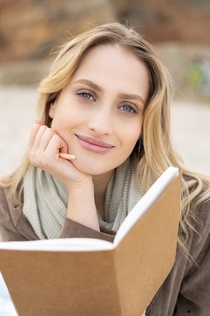 Foto retrato de una hermosa joven sosteniendo un cuaderno abierto con una leve sonrisa sentado en una playa