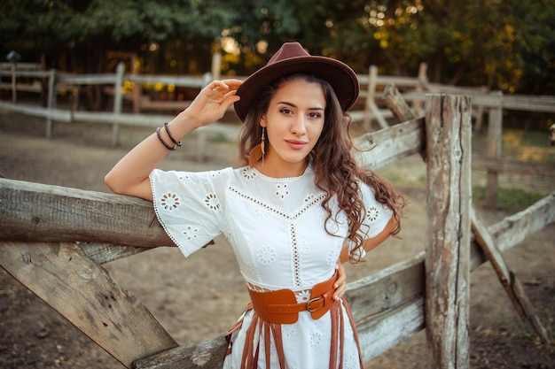 Retrato de una hermosa joven con un vestido blanco con botas sobre la rodilla y un sombrero con cinturón y flecos en un establo rústico