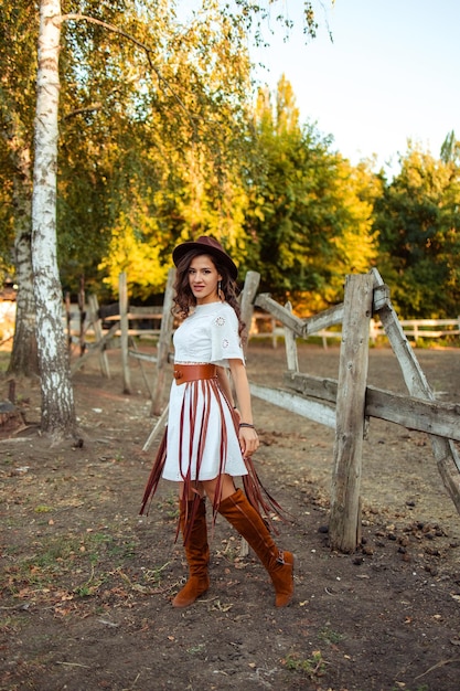 Retrato de una hermosa joven con un vestido blanco con botas sobre la rodilla y un sombrero con cinturón y flecos en un establo rústico