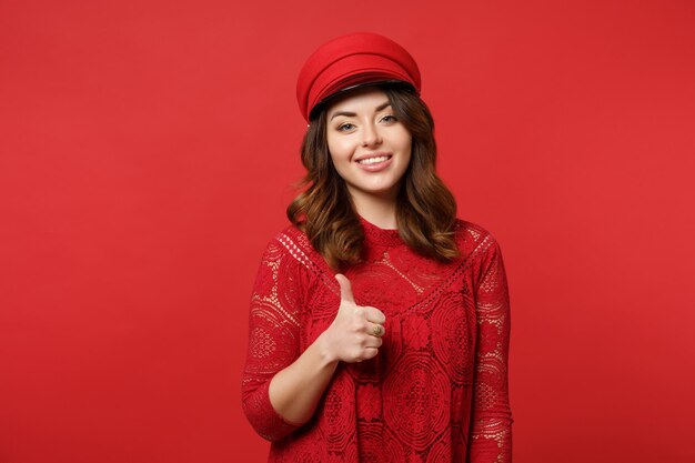 Retrato de una hermosa joven con vestido de encaje y cámara con aspecto de gorra que muestra el pulgar hacia arriba aislado en un fondo de pared rojo brillante en el estudio. Gente emociones sinceras concepto de estilo de vida. Simulacros de espacio de copia.