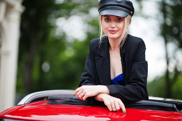 Retrato de una hermosa modelo rubia de moda sexy con gorra y todo negro con maquillaje brillante en el techo corredizo abierto del coche rojo de la ciudad