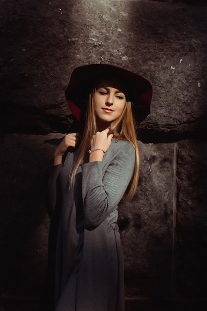 Retrato de hermosa modelo rubia con vestido gris cálido y sombrero rojo. Mujer posando en la luz del sol y la sombra en la calle