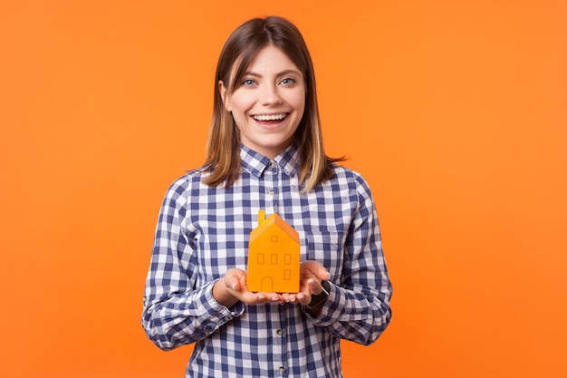 Retrato de una hermosa morena encantada con una sonrisa encantadora usando una camisa a cuadros de pie sosteniendo una casa de papel en las manos y mirando la cámara en un estudio interior aislado en un fondo naranja