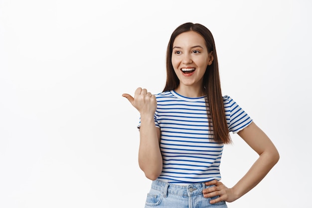 Retrato de una hermosa morena riéndose, sonriendo feliz y señalando mirando a la izquierda a la venta, mostrando descuentos, pancarta de la tienda en el espacio de la copia, de pie sobre fondo blanco.