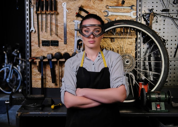 Retrato de una hermosa morena con ropa de trabajo, delantal y gafas, de pie con los brazos cruzados en un taller contra herramientas de pared.