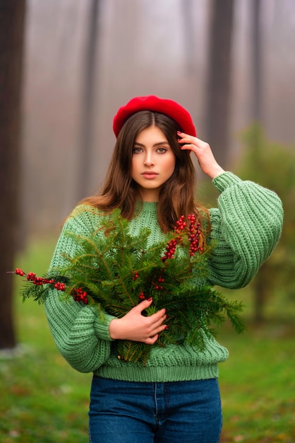 Retrato de una hermosa morena con sombrero rojo y suéter de punto verde sosteniendo ramas navideñas