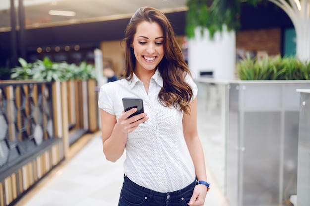 Retrato de hermosa morena sonriente en camisa con teléfono inteligente en el centro comercial.