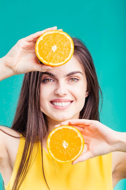 Retrato de una hermosa morena sonriente sosteniendo dos partes de fruta naranja cortadas cerca de su cara antes de un fondo colorido concepto de frescura saludable