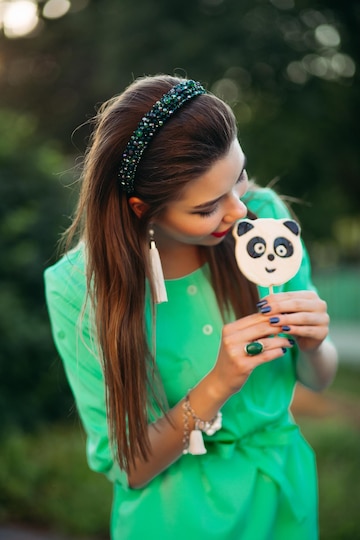 Retrato de una hermosa morena con vestido verde, comiendo dulces como panda en un palo y mirando al costado. mujer bonita y elegante con labios rojos, aretes largos y banda para el Retrato de una hermosa morena con vestido verde, comiendo dulces como panda en un palo y mirando al costado. mujer bonita y elegante con labios rojos, aretes largos y banda para el