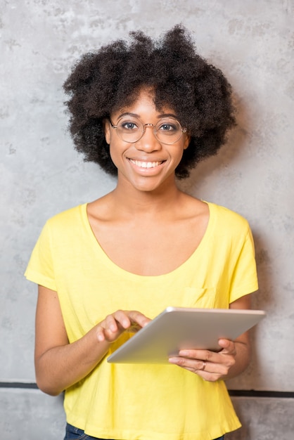 Retrato de una hermosa mujer africana en camiseta amarilla con tableta digital cerca de la pared gris en el interior