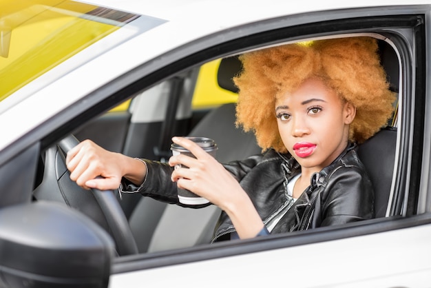 Retrato de una hermosa mujer africana en chaqueta de cuero con taza de café sentado en el coche sobre el fondo amarillo