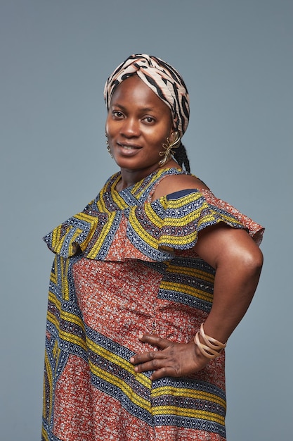 Retrato de hermosa mujer africana en traje nacional sonriendo a la cámara de pie contra la espalda azul