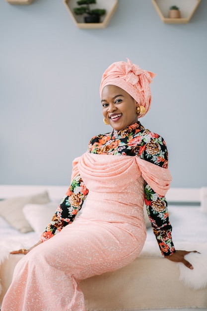 Retrato de hermosa mujer afroamericana con maquillaje de moda y joyería, con vestido rosa étnico y pañuelo en la cabeza, posando en la cámara con una sonrisa sentada en la cama en el estudio.