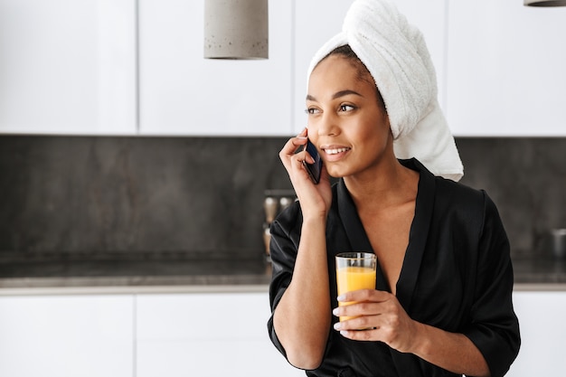 Retrato de hermosa mujer afroamericana vistiendo bata, a través de teléfono móvil en la cocina