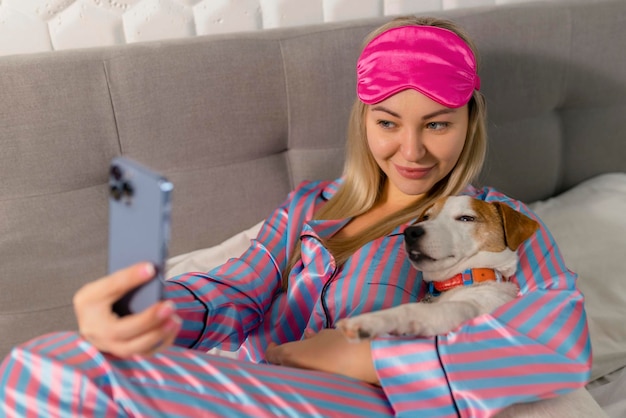 Retrato de una hermosa mujer alegre en pijama abrazando a un perro usando un teléfono móvil para una selfie sonriendo mientras se acuesta en la cama después de dormir o tomar una siesta