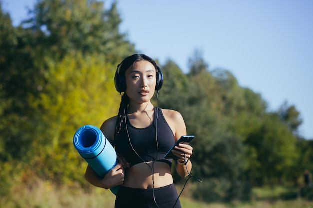 Retrato de una hermosa mujer asiática delgada, atleta en el bosque, escuchando música con auriculares y sosteniendo una alfombra de yoga, atleta sonriendo y mirando la cámara