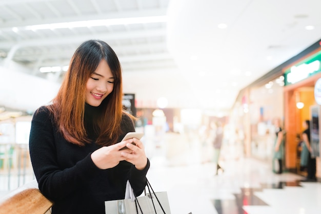 Retrato de hermosa mujer asiática joven en centro comercial, sonriendo con teléfono inteligente