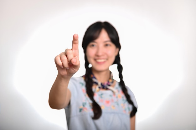 Retrato de hermosa mujer asiática joven feliz en vestido de mezclilla tocando pantalla invisible sobre fondo blanco