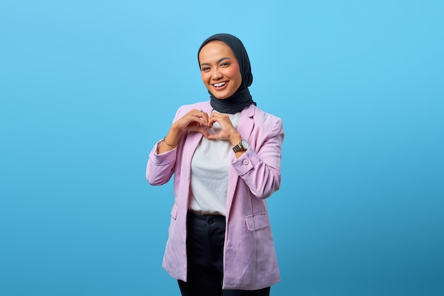 Retrato de hermosa mujer asiática mostrando gesto de forma de corazón sobre fondo azul.