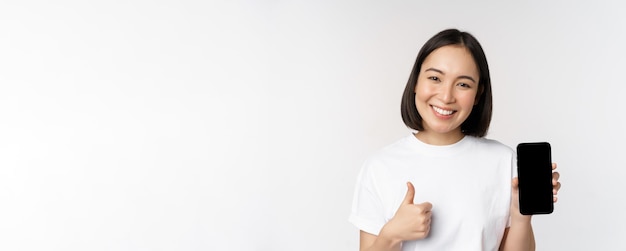 Retrato de una hermosa mujer asiática que muestra el pulgar hacia arriba y la pantalla del teléfono inteligente sonriendo mientras anuncia m