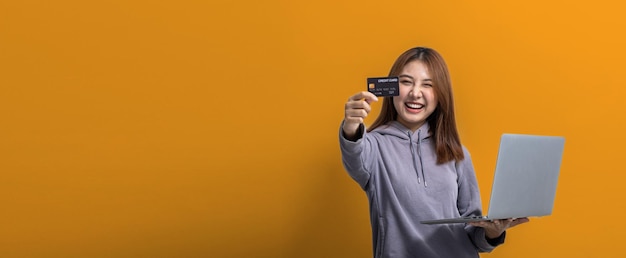 Retrato de una hermosa mujer asiática que sostiene una tarjeta de crédito y una computadora portátil en un concepto de retrato de fondo aislado utilizado para publicidad y señalización aislado sobre un espacio de copia de fondo amarillo