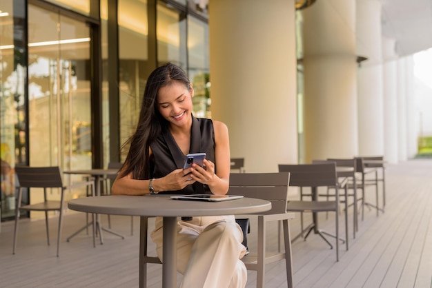 Retrato de hermosa mujer asiática sentada al aire libre en el restaurante cafetería sonriendo y usando el teléfono