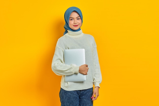 Retrato de una hermosa mujer asiática sonriente con suéter blanco y hiyab sosteniendo una computadora portátil y mirando a la cámara aislada sobre el concepto de estilo de vida religioso de la gente de fondo amarillo