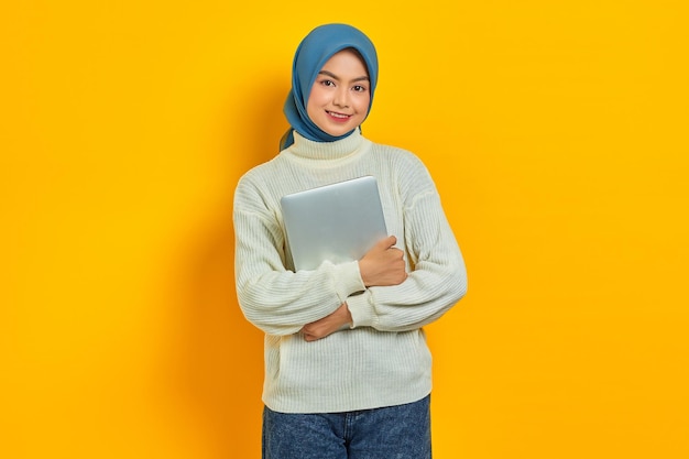 Retrato de una hermosa mujer asiática sonriente con suéter blanco y hiyab sosteniendo una computadora portátil y mirando a la cámara aislada sobre el concepto de estilo de vida religioso de la gente de fondo amarillo