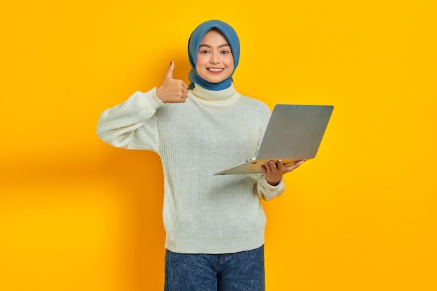 Retrato de una hermosa mujer asiática sonriente con suéter blanco y hiyab sosteniendo una computadora portátil que muestra el pulgar hacia arriba aislado sobre fondo amarillo Concepto de estilo de vida religioso de la gente